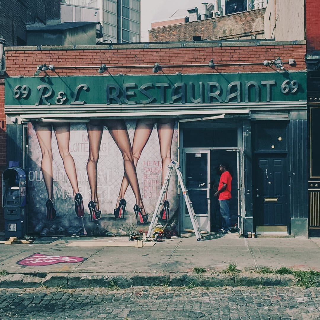 The storefront of a restaurant with a mural of women's legs in stockings and high heels painted on the wall. The sign reads 'R & L RESTAURANT 69'. There are steps, a ladder, and a man in a red shirt near the entrance.