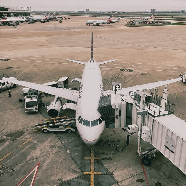 View of an airplane at an airport gate connected to a jet bridge, with luggage carts and ground crew vehicles nearby, and other airplanes parked on the tarmac in the background.