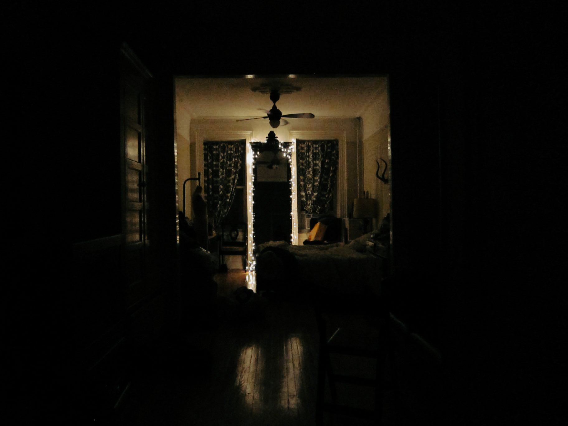 A dark room with a view of a lit bedroom in the distance, decorated with string lights and patterned curtains.