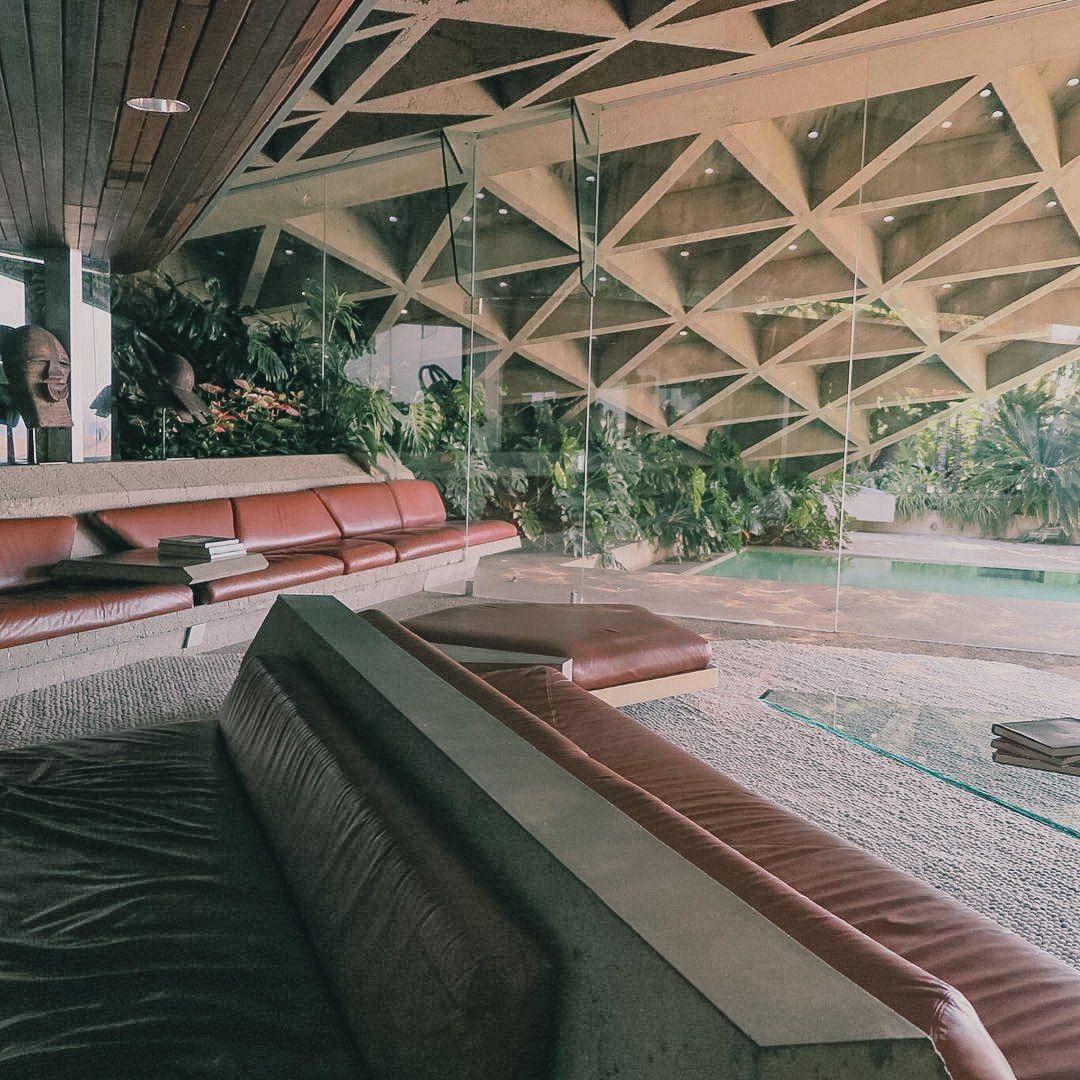 A modern indoor lounge area with leather seating, glass walls, greenery, and a view of a swimming pool outside.