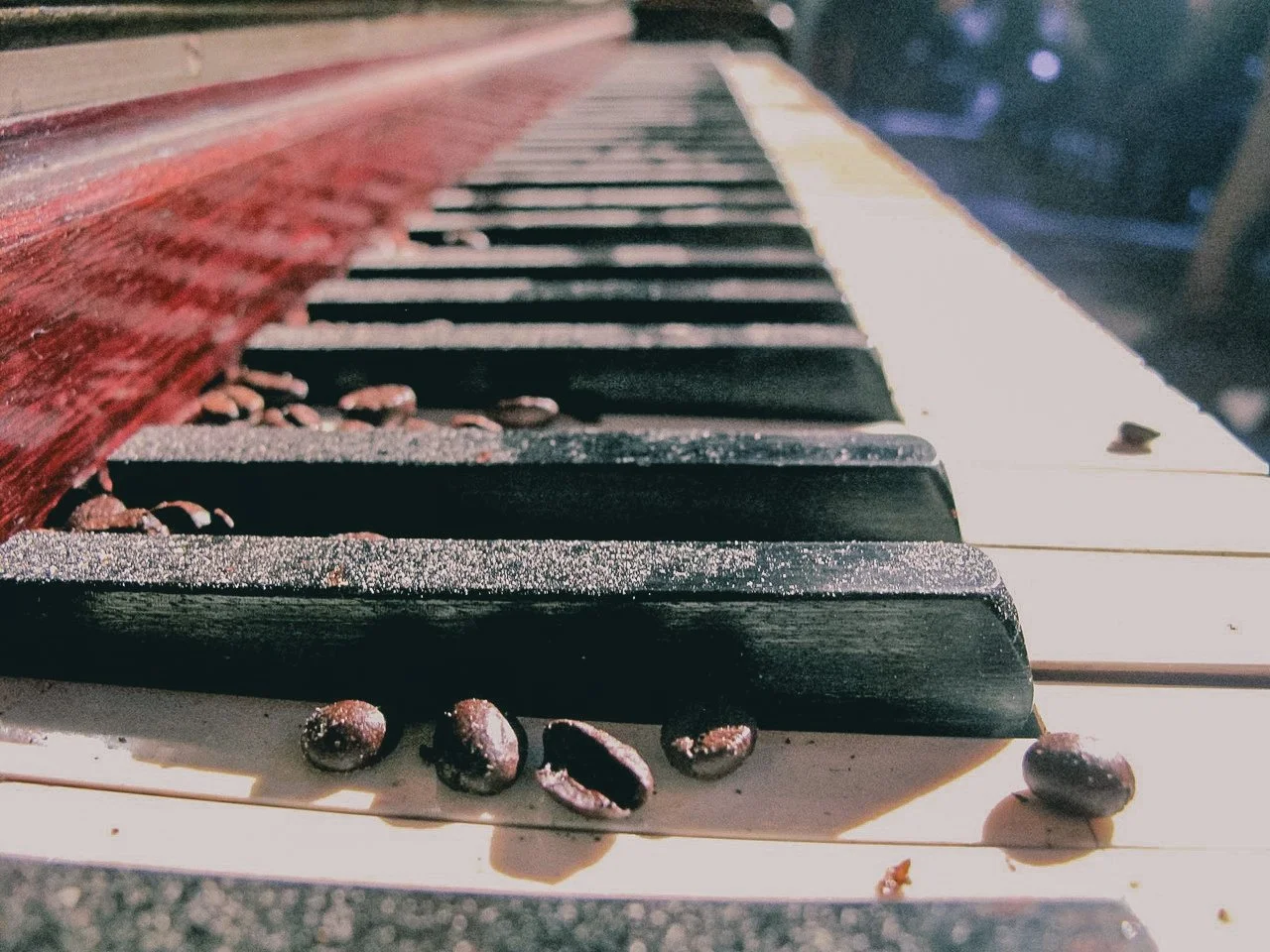 Close-up of a dusty piano keyboard with coffee beans scattered on and around the keys, with a red wooden part of the piano visible on the left.