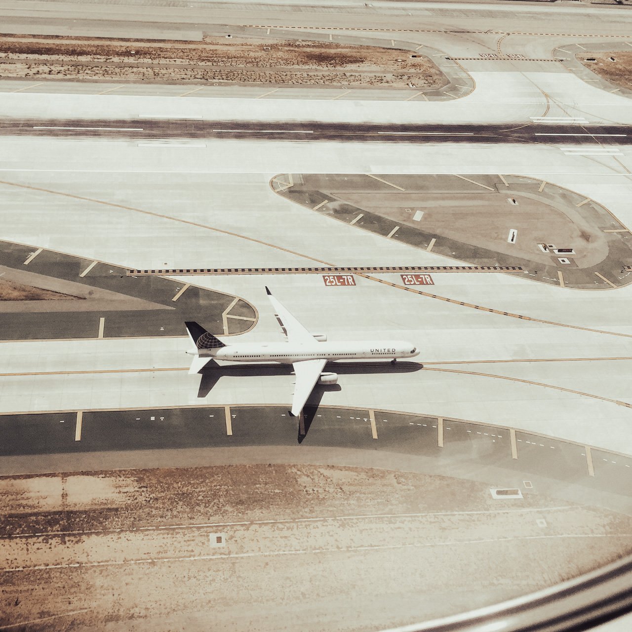 An airplane parked on a tarmac seen from a window, with markings and signs on the ground, and brown soil surrounding the paved area.