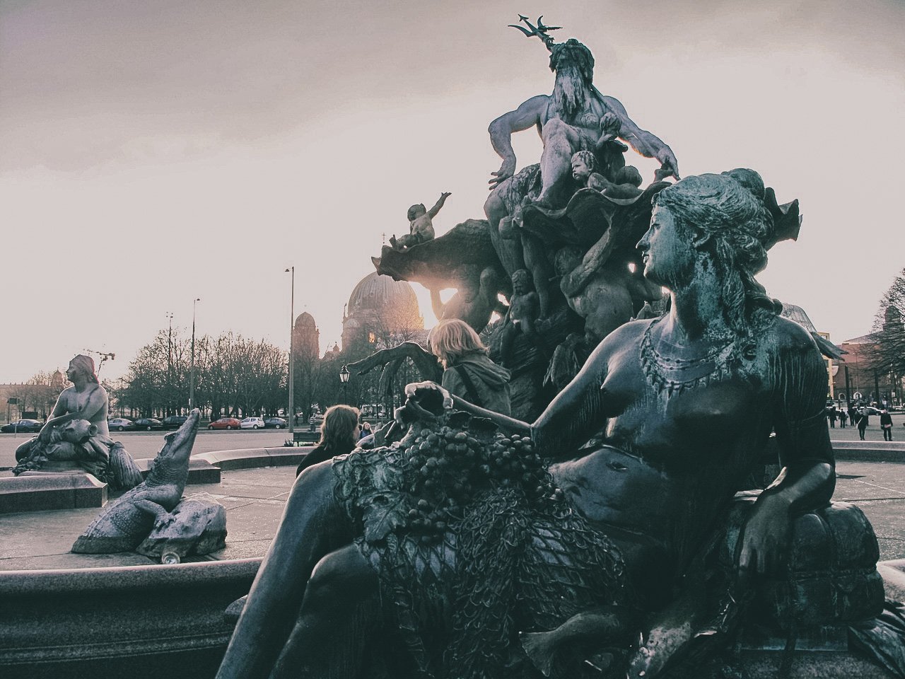 Bronze fountain sculpture with multiple mythological figures, including a mermaid, a woman with a fish tail, and children in an outdoor setting with trees and cars in the background, under an overcast sky.