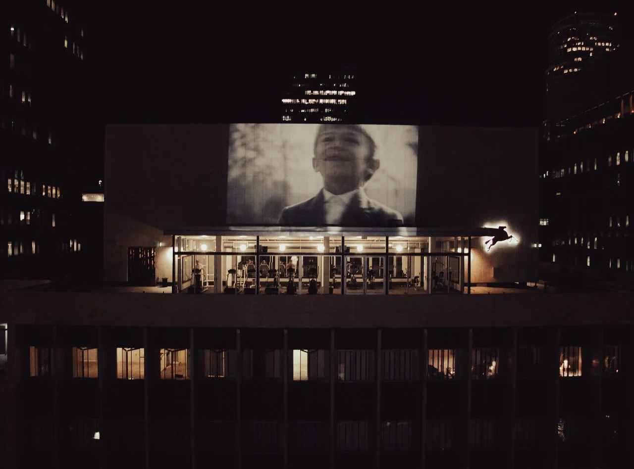 Nighttime cityscape with a large outdoor movie screen displaying a black-and-white image of a smiling boy in a suit. The building has multiple floors with illuminated rooms and a gym area with visible equipment on the rooftop, where a person is seen 