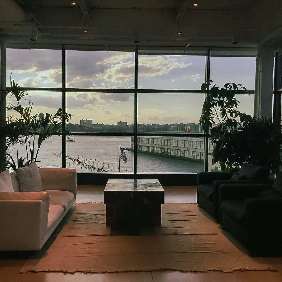 Interior of a modern room with large floor-to-ceiling windows overlooking a body of water, with a cityscape in the background. The room has a white sofa on the left, black armchairs on the right, a wooden coffee table in the center, and several indoo