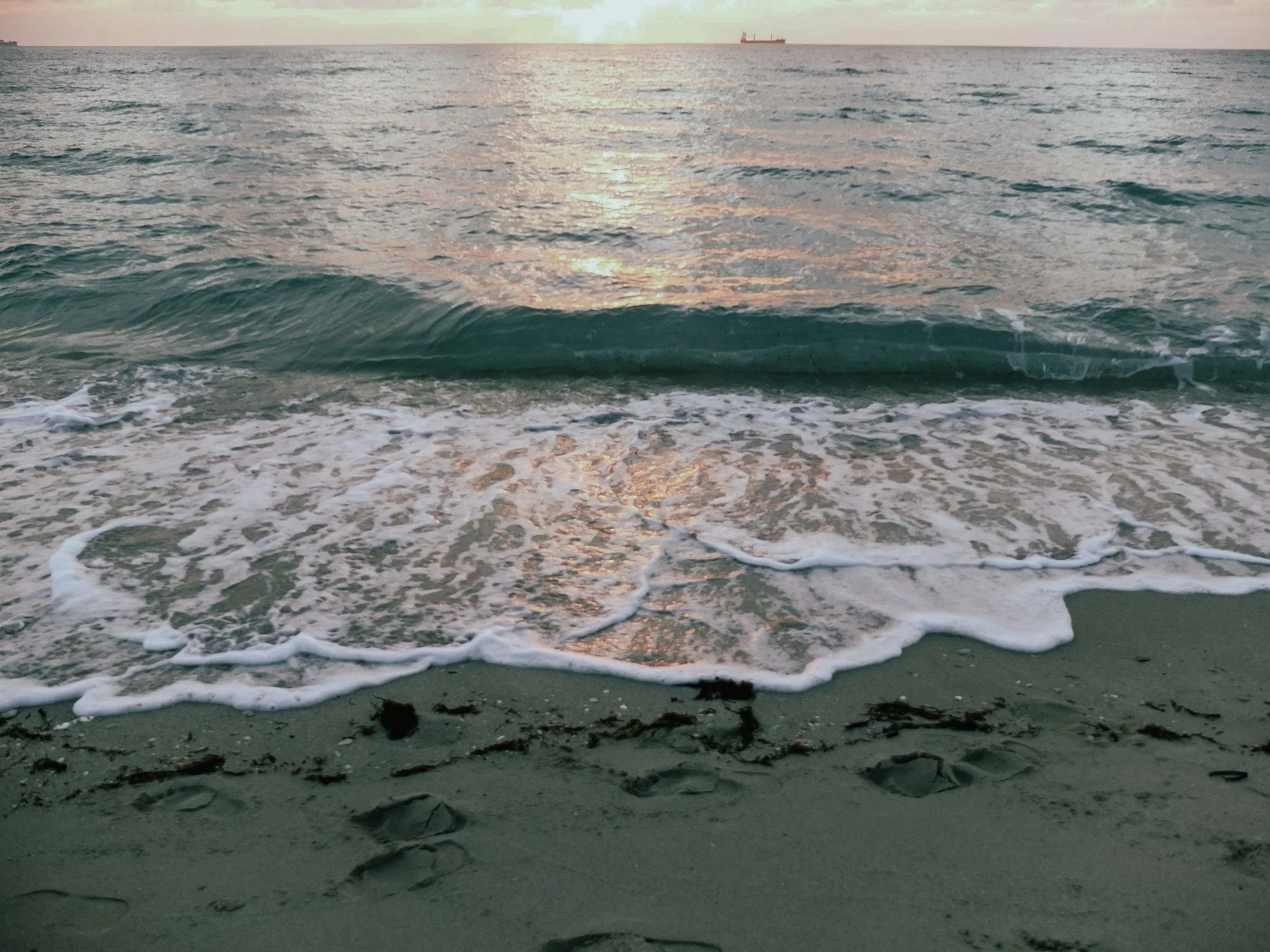 Sunset over the ocean with waves crashing onto sandy beach, ship in the distance.