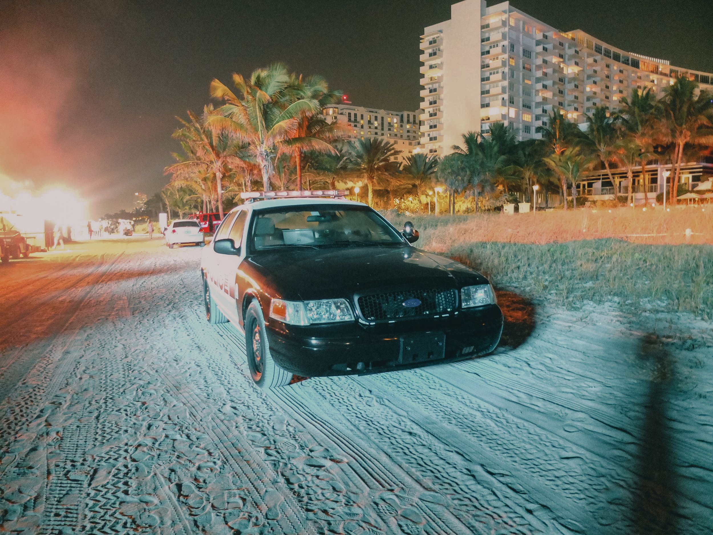 Nighttime scene on a sandy beach shows a black police car parked near palm trees and a large hotel or apartment building in the background. There are other vehicles and bright lights illuminating the area.