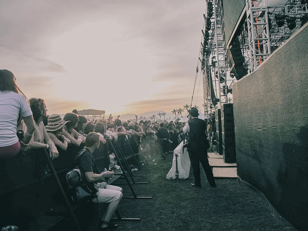 Crowd waiting for a concert to start at sunset, with a stage set up on the right and palm trees in the distance.
