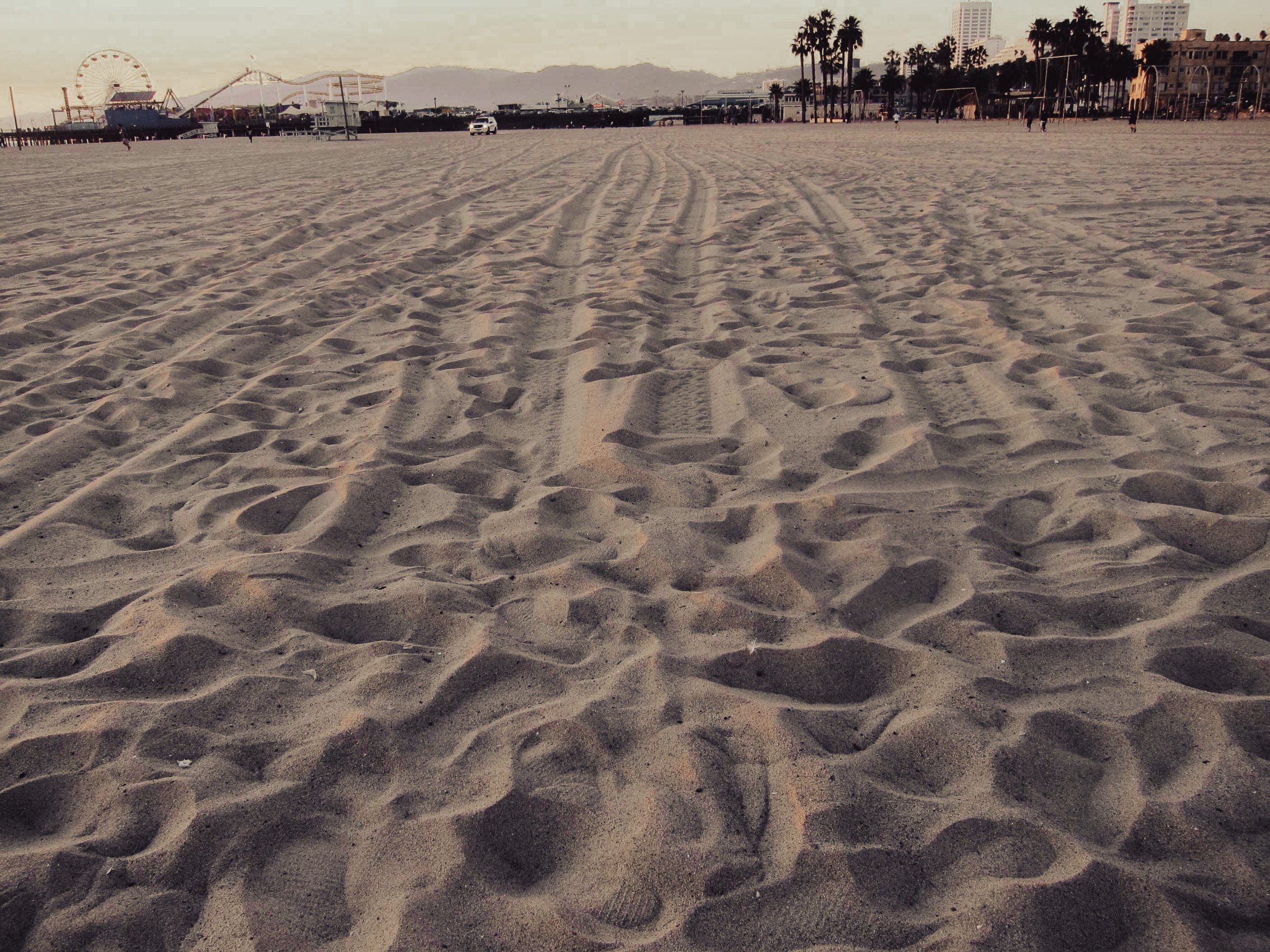 Empty sandy beach with tire tracks leading toward the carnival and skyline in the background, with palm trees and park area.