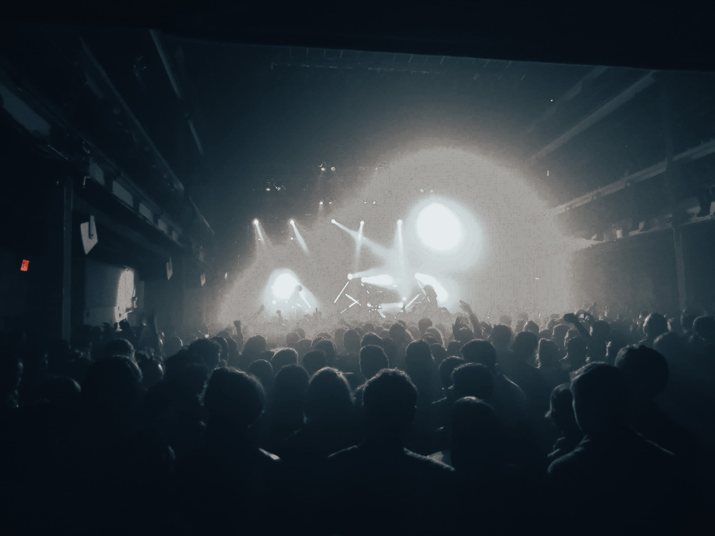 People attending a concert or live performance in a dark indoor venue, with stage lights shining brightly and silhouettes of the audience visible.
