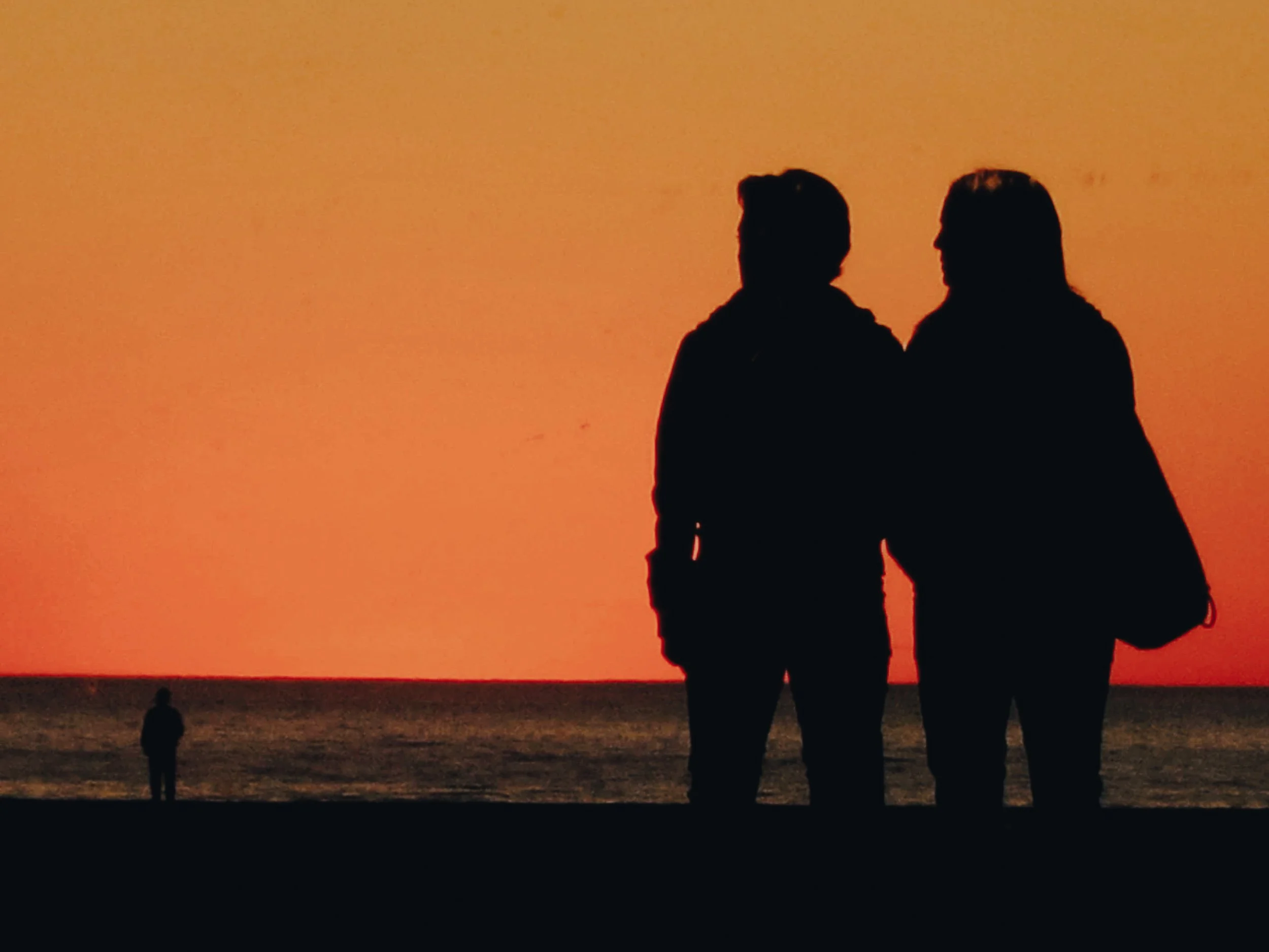 Silhouettes of a man and woman standing close together on a beach during sunset, with a third person in the distance near the water.