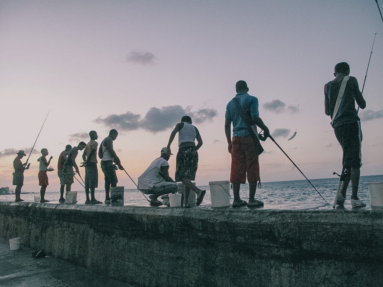 People fishing on a pier during sunset or dawn, with some holding fishing rods and one person sitting near buckets by the water.