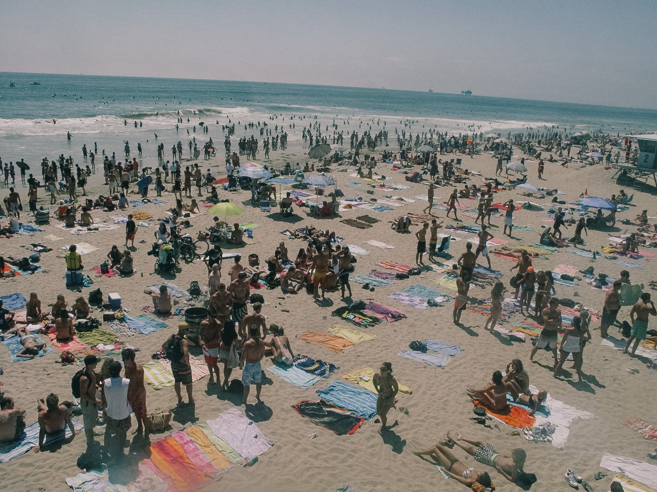 Crowded beach with many people sunbathing, walking, and playing near the shoreline on a sunny day.