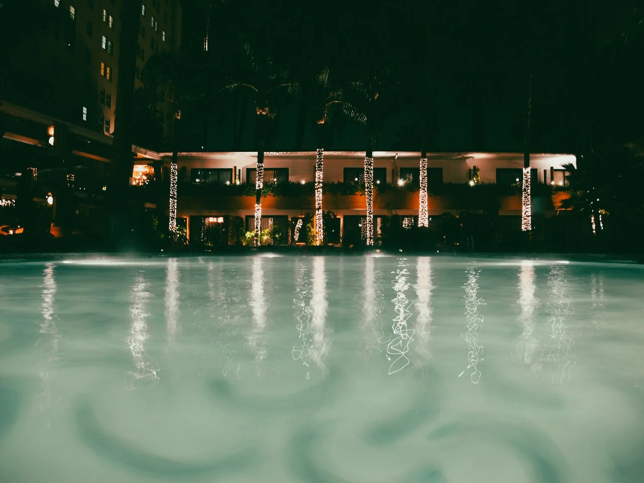 Night view of a pool with the hotel or building in the background, illuminated with decorative lights wrapped around the trees.