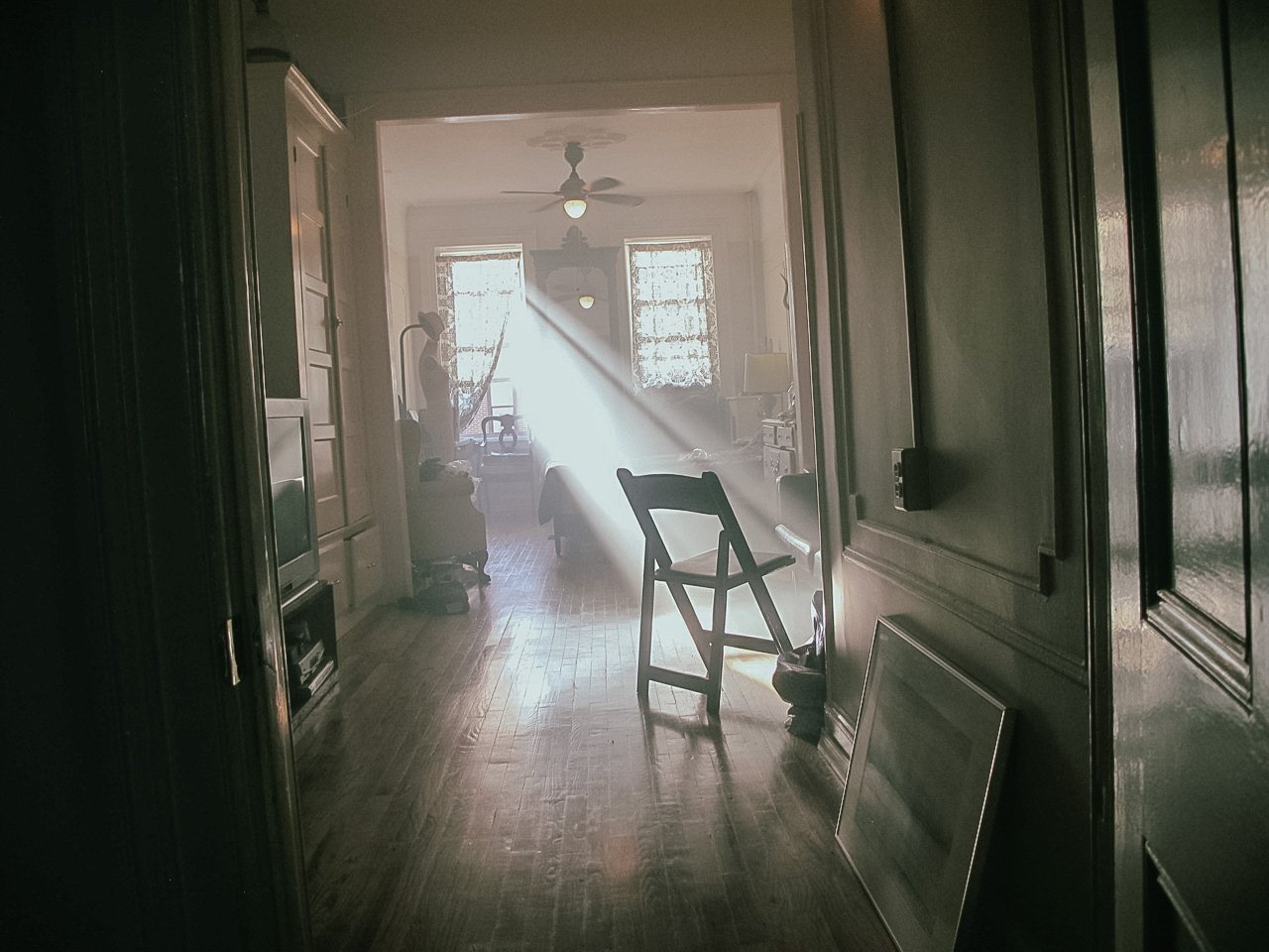 A living room with sunlight streaming through a window, featuring a ceiling fan, a chair, and various furniture and objects, with a wooden floor and a doorway leading to another room.
