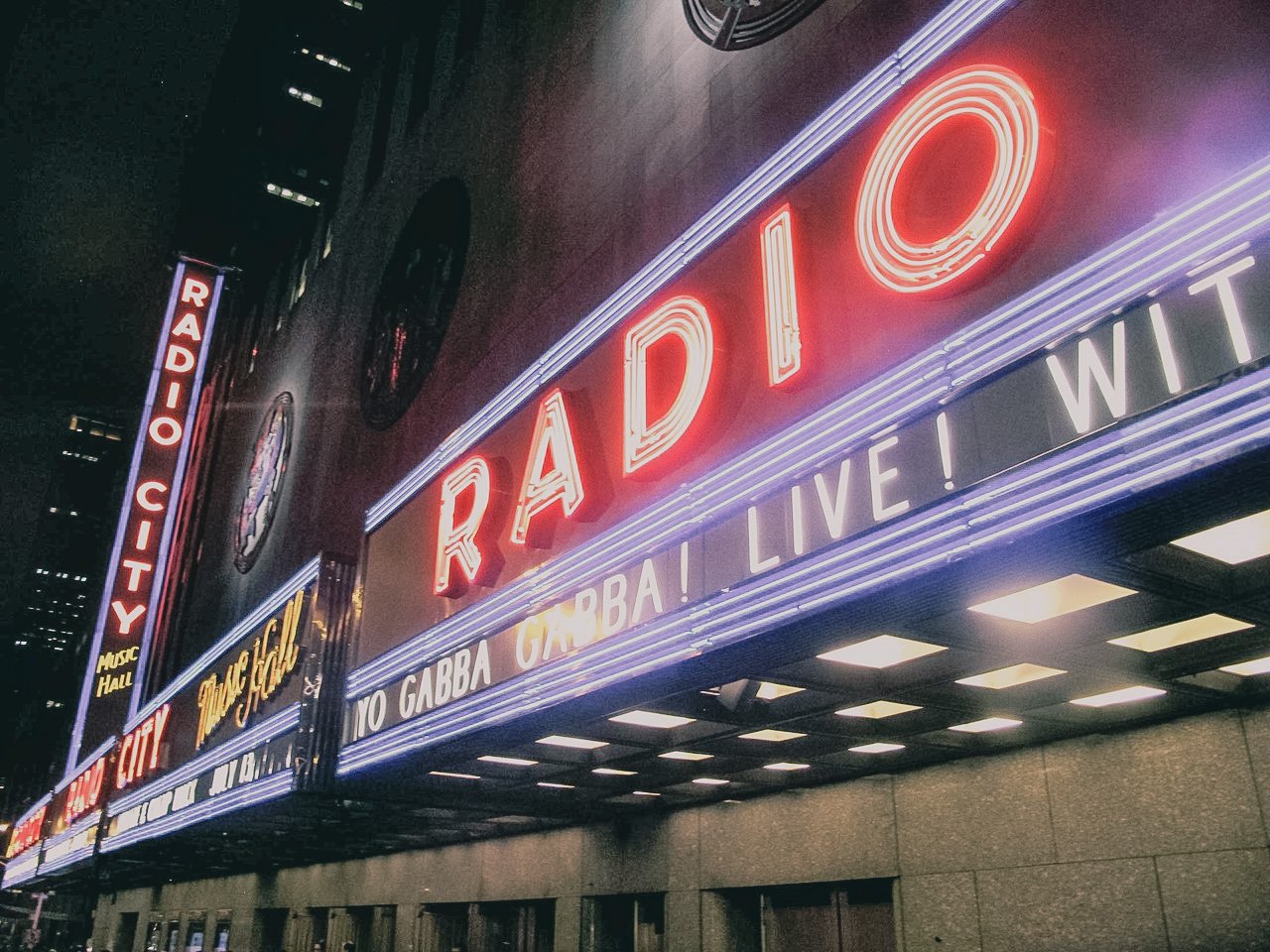 Night view of Radio City Music Hall marquee with bright neon lights displaying the words "RADIO CITY" and phrases such as "TOO GABBA GABBA! LIVE! WIT" on the marquee.