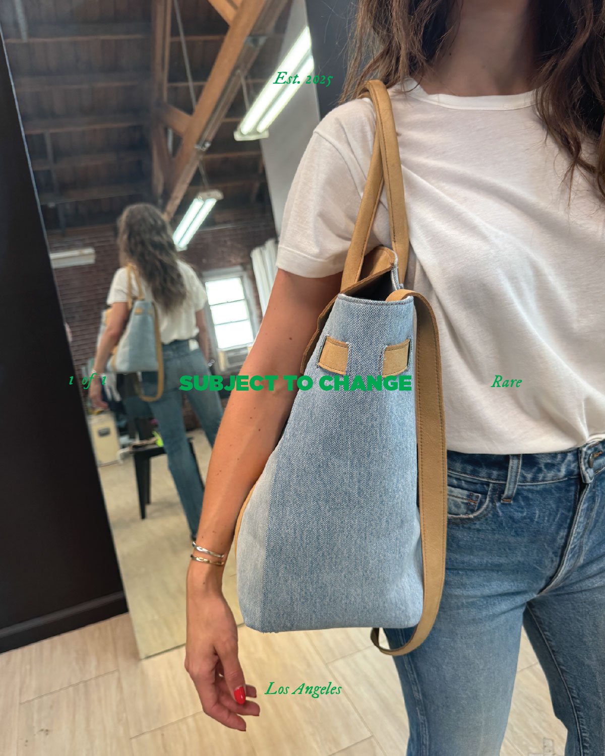 Woman in a white t-shirt and blue jeans standing in front of a mirror in a store, holding a light blue denim tote bag with tan straps over her shoulder. The store has a rustic interior with exposed brick and wooden beams, and a window in the backgrou