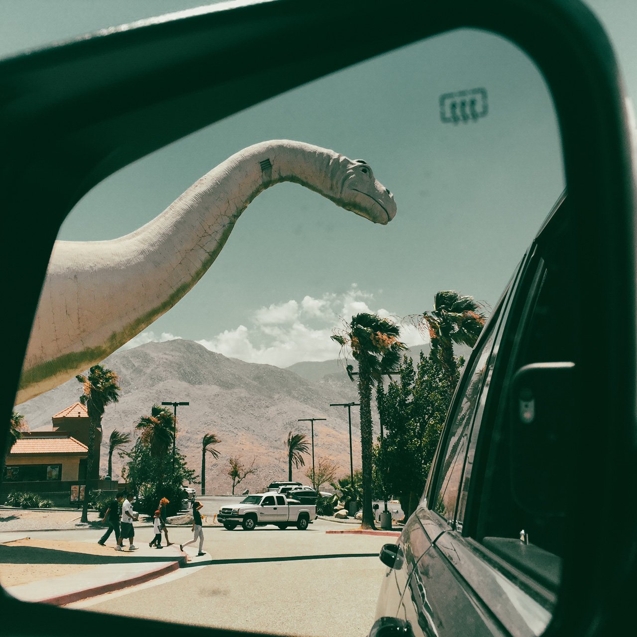 A large dinosaur sculpture peeking over trees and a parking lot, seen through a vehicle's side mirror.