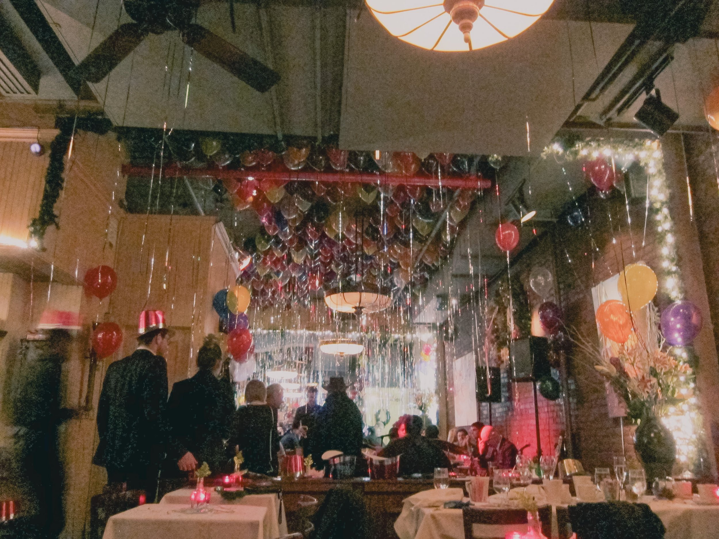A cozy indoor restaurant or party venue decorated with lots of colorful balloons and string lights. Several people are gathered, some are sitting at tables while others are standing, possibly celebrating an event like a birthday. Decor includes a lar