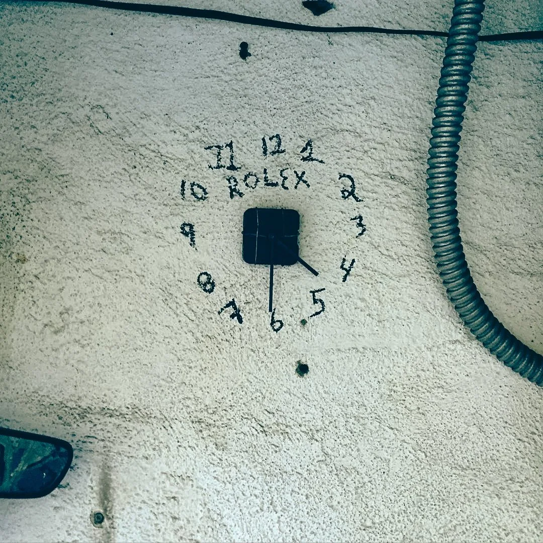 Clock painted on a textured wall with numbers arranged in a circle and the word 'ROLEX' in the center, with a black switch or control in the middle and a wire running along the wall.