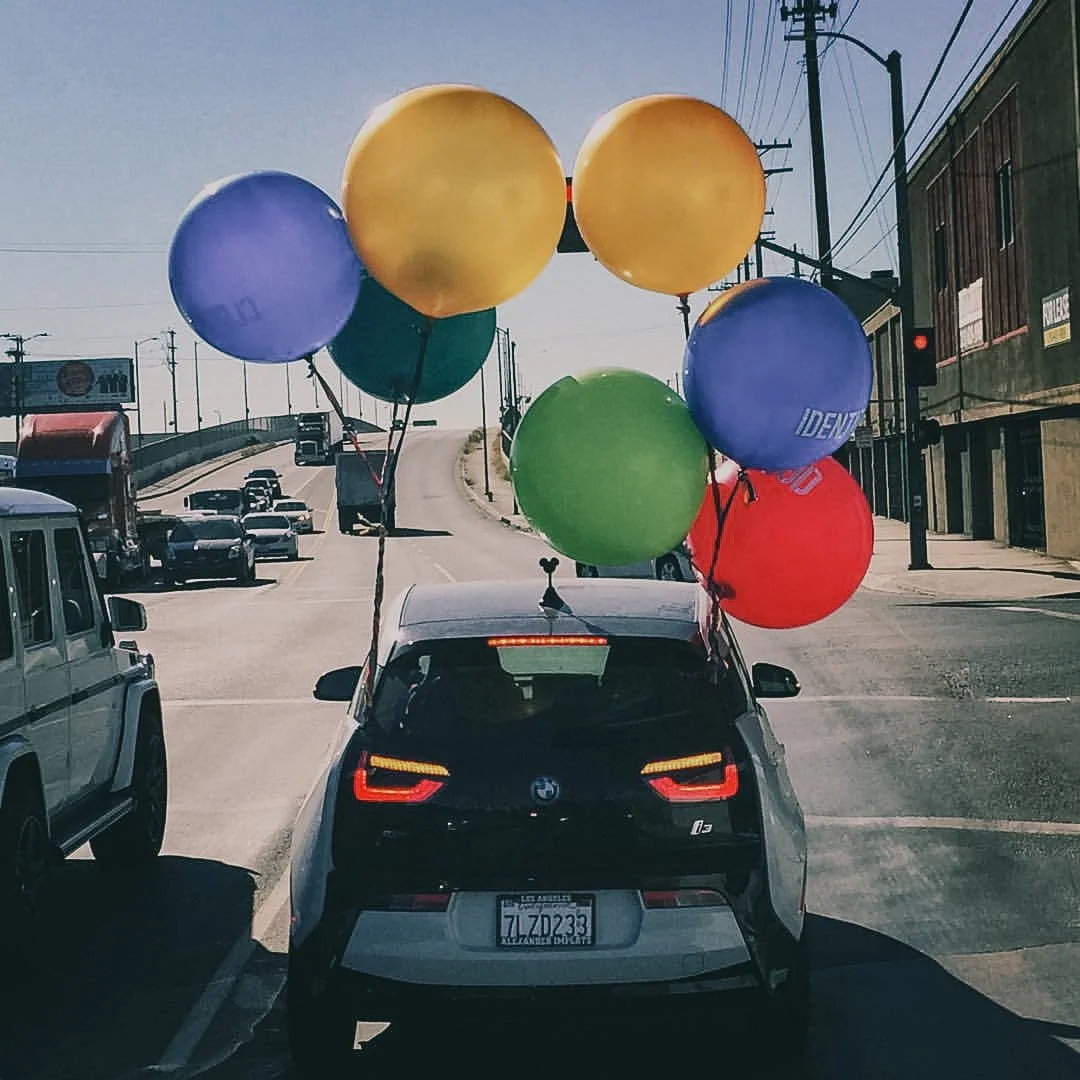 A black BMW i3 car with colorful balloons attached to its roof driving on a street in an urban area.