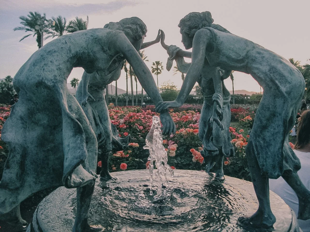Bronze sculpture of two children playing near a small fountain with water, set against a garden with pink flowers and palm trees.