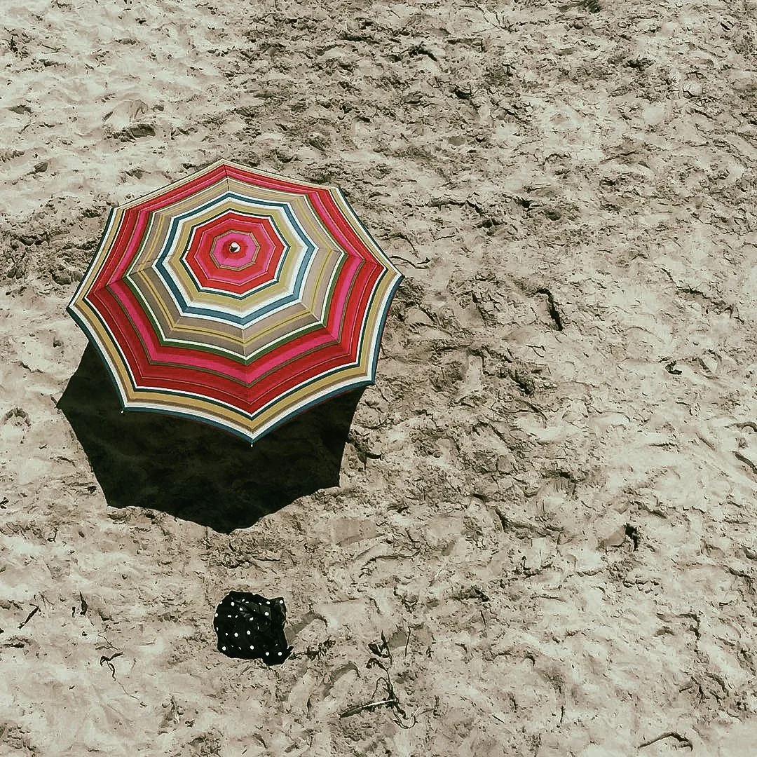 A colorful striped beach umbrella casting a shadow on the sandy beach, with a small black and white polka dot cloth or bag nearby.