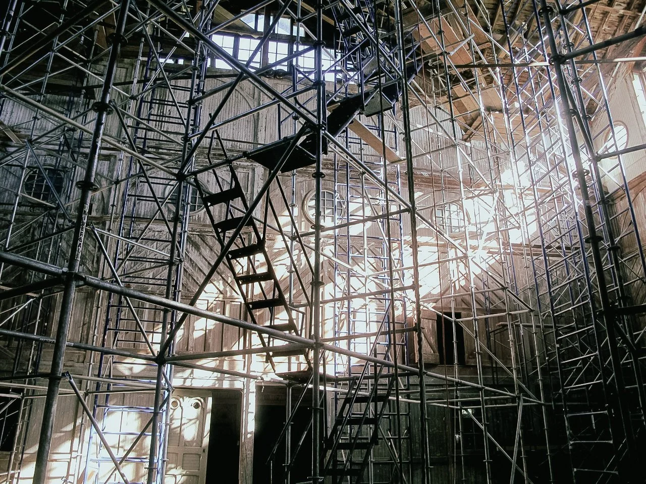 Interior of an old building with extensive metal scaffolding and wooden stairs, sunlight streaming through windows.