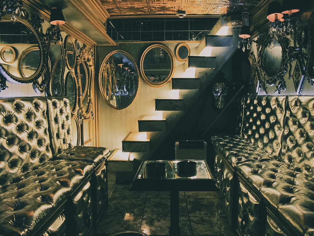 Luxurious interior with black tufted leather sofas, ornate gold-framed mirrors on the wall, and a staircase with a black railing leading upward. Dim lighting and gold accents give a glamorous vibe.