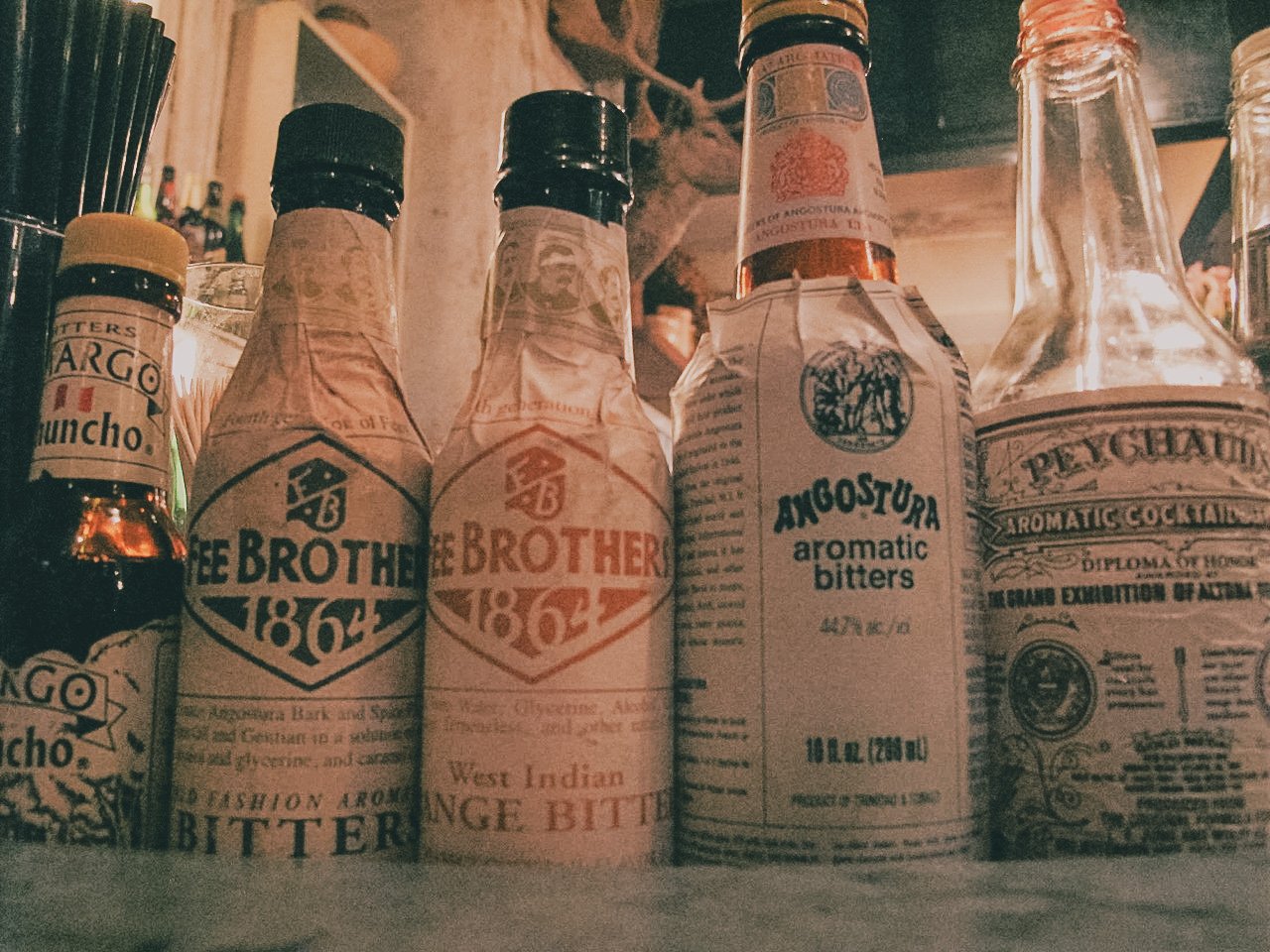 A row of vintage cocktail bottles on a bar shelf, including Angostura aromatic bitters and other bitter liqueurs, with some bottles wrapped in paper labels and others with clear glass.
