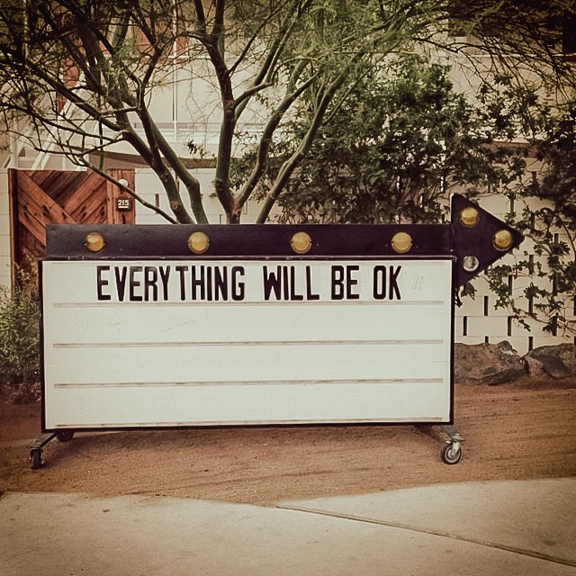 A white message board with black border and wheels displaying the text "EVERYTHING WILL BE OK". The board is outdoors on a dirt surface, with trees and a building with a wooden fence in the background. There is an arrow-shaped sign on the right side 