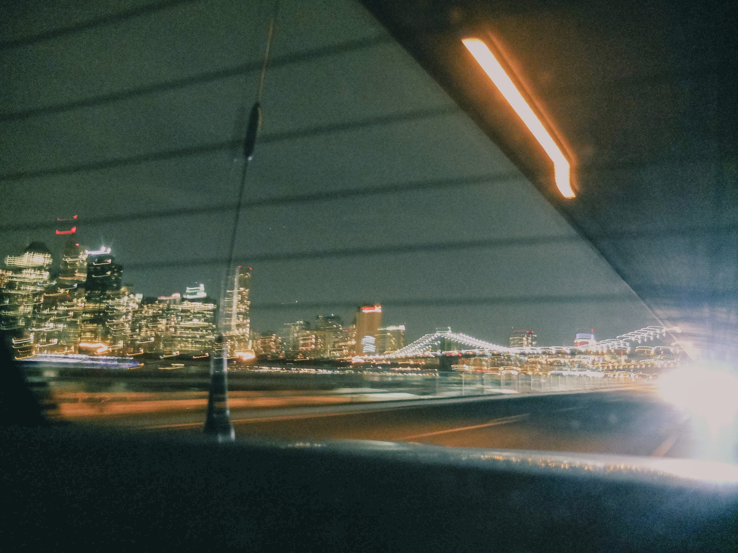 Nighttime city skyline view through a window with horizontal blinds, with blurred city lights and a bridge illuminated in the distance.