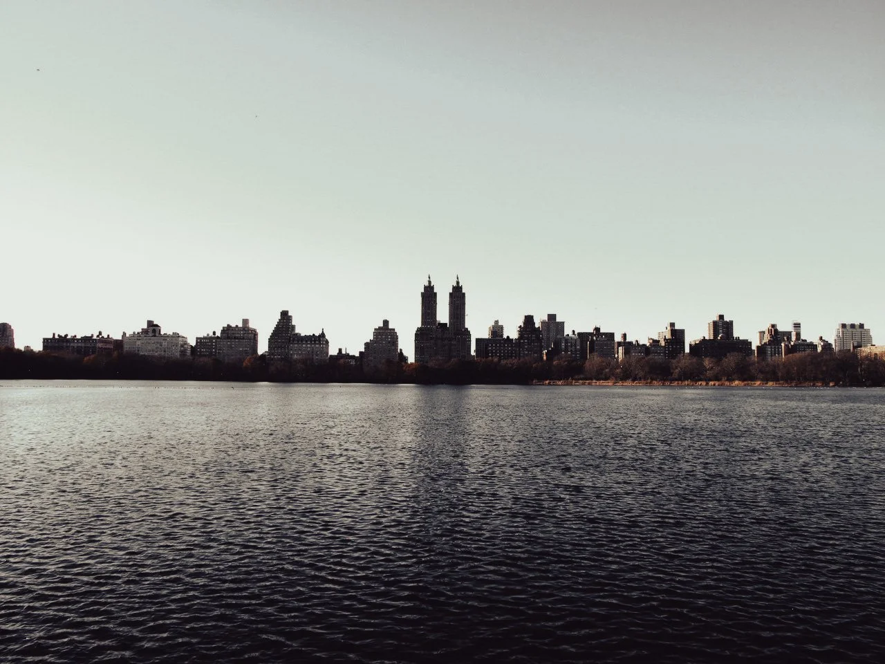 City skyline with tall buildings across a body of water with a calm surface, trees along the shoreline, under a clear sky.