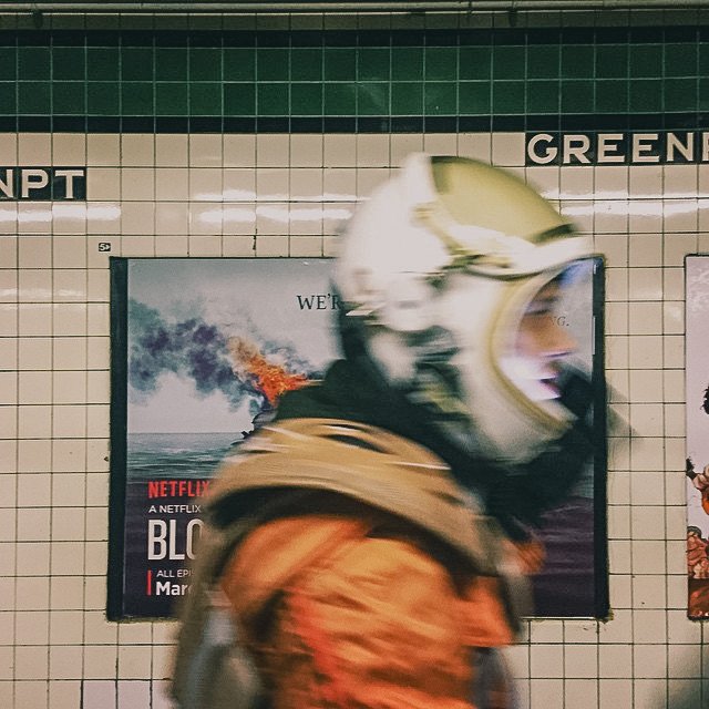 A person in a motorcycle helmet standing at a subway station with a green tiled wall and a Netflix advertisement poster in the background.