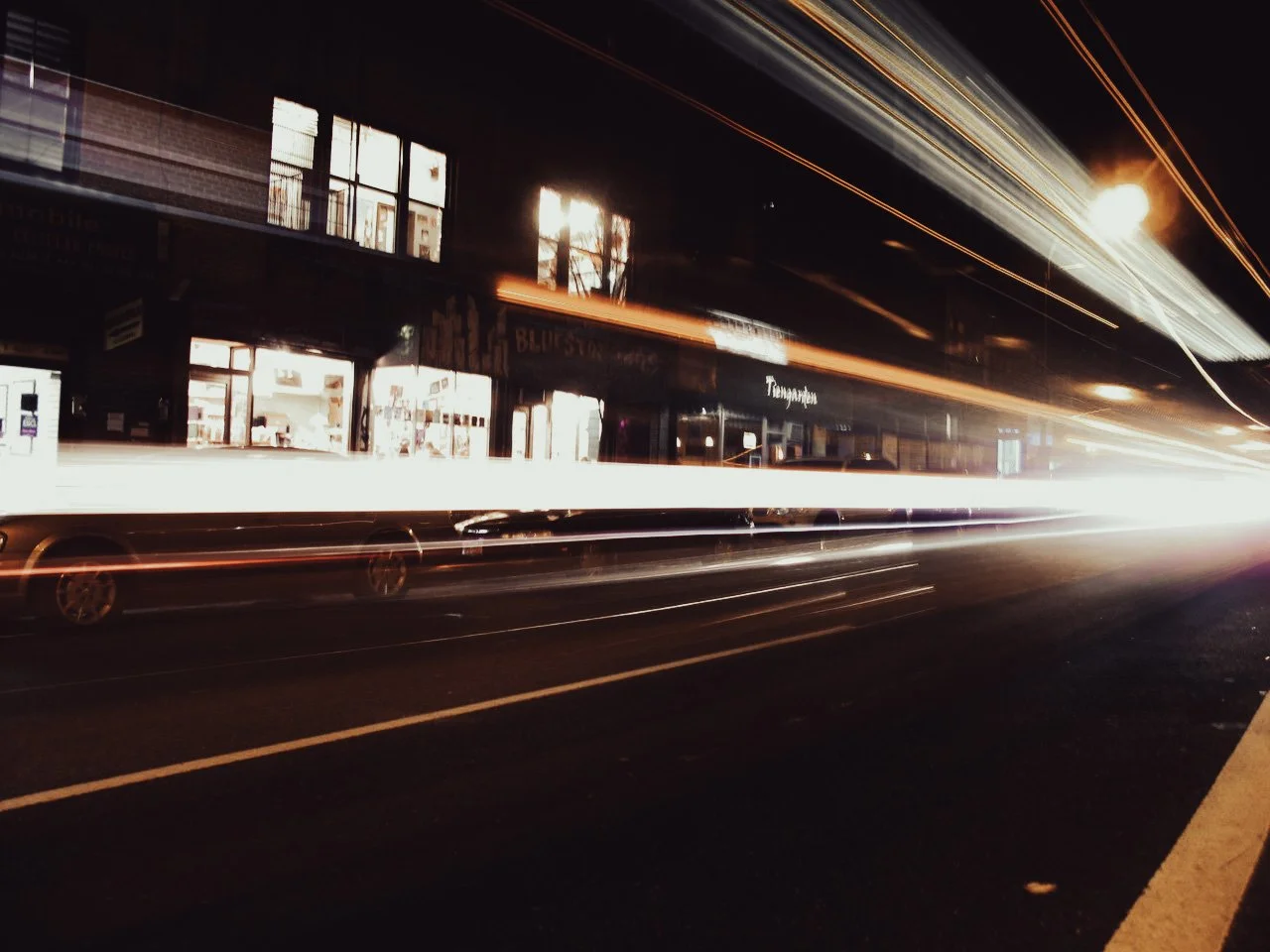 Nighttime city street scene with blurred moving cars and light streaks from headlights and taillights, storefronts with lit windows, and reflections on the glass.