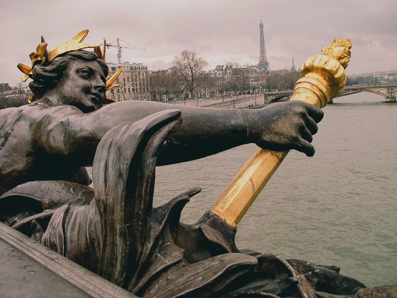 Close-up of the statue of a river young woman holding a torch in Paris, with the Eiffel Tower and a bridge over the Seine river in the background on a cloudy day.