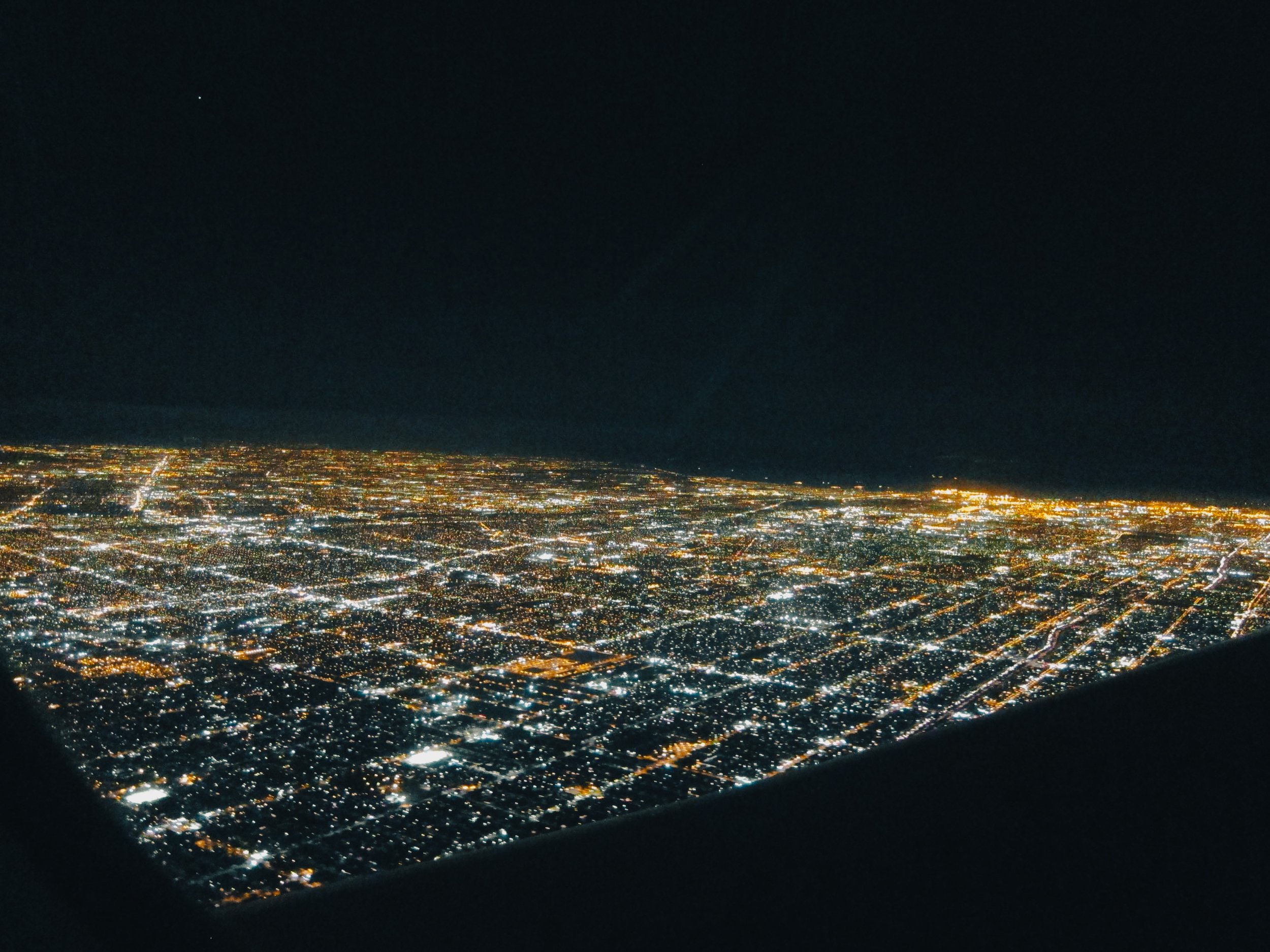 Nighttime aerial view of a city with brightly lit streets and buildings, seen from an aircraft window.