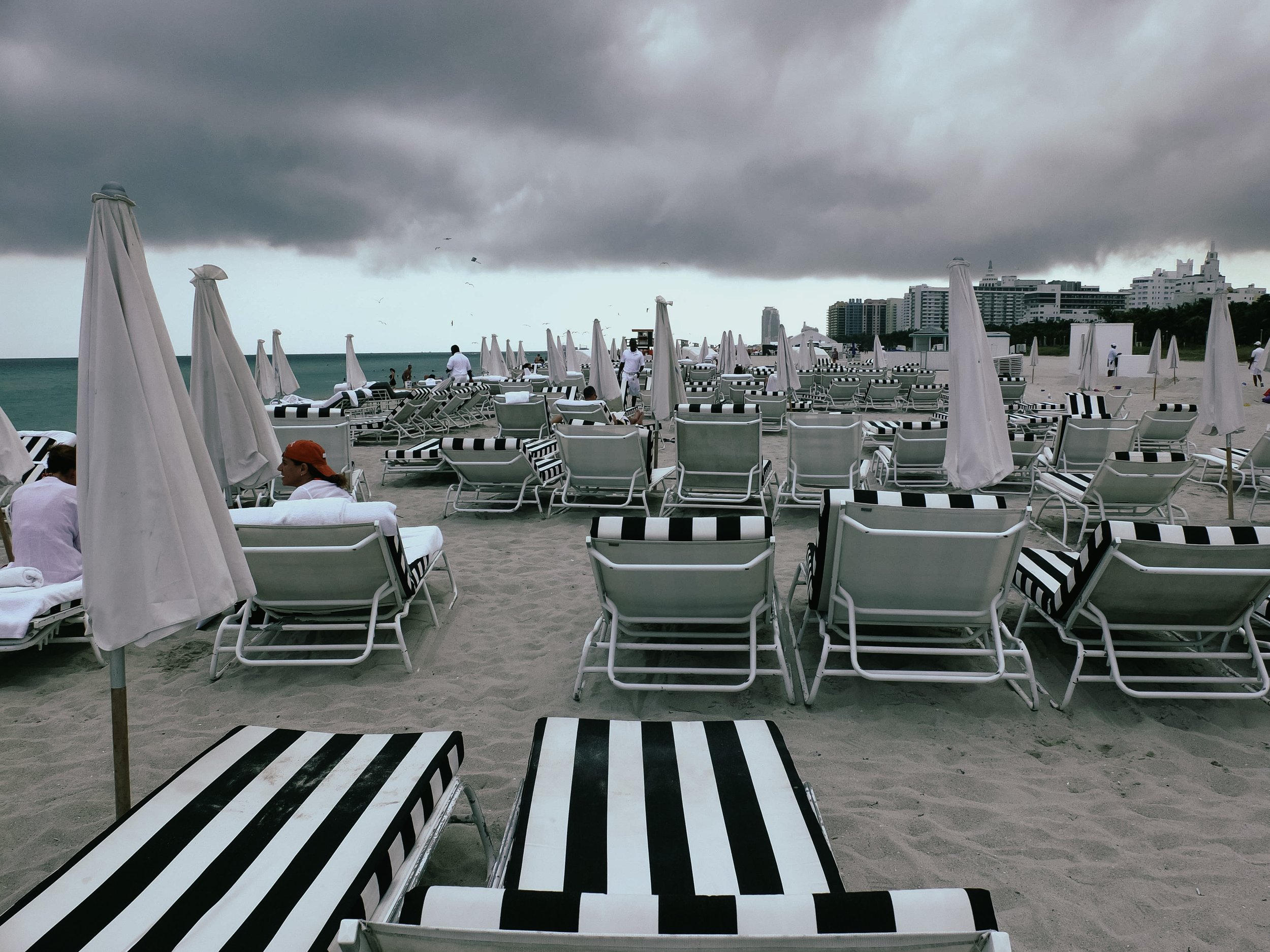 Overcast beach with numerous white lounge chairs and umbrellas, some people sitting or walking, and city buildings in the distance.