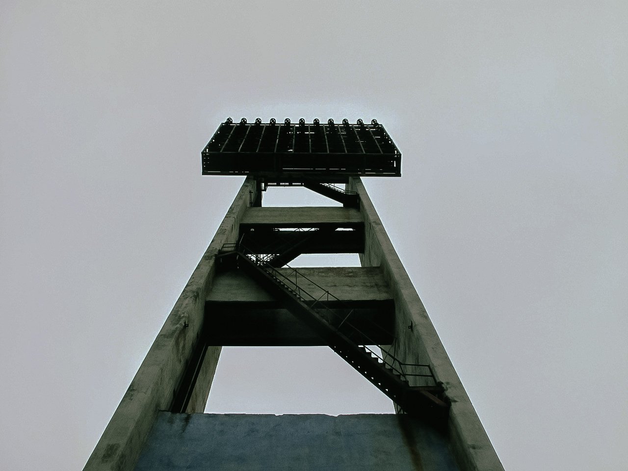 Low-angle view of a tall concrete tower with a metal platform at the top, set against an overcast sky.