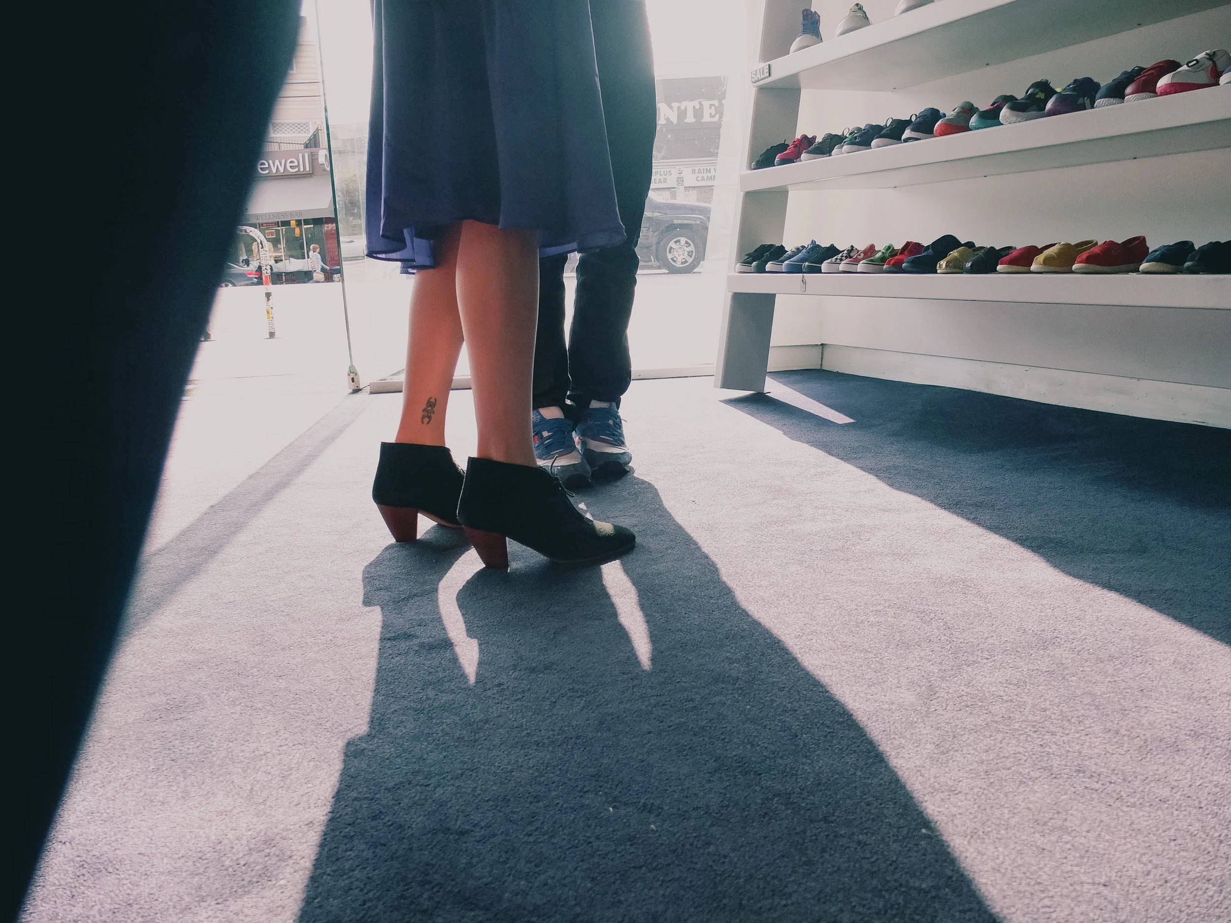 A woman trying on black high heels inside a shoe store, with a man standing nearby. Shoe shelves filled with colorful sneakers are visible on the right side. Sunlight streams through the glass door at the front of the store.