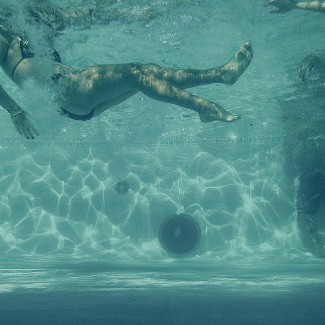 An underwater photo of a person swimming horizontally in a pool, with sunlight creating ripples on the pool floor.
