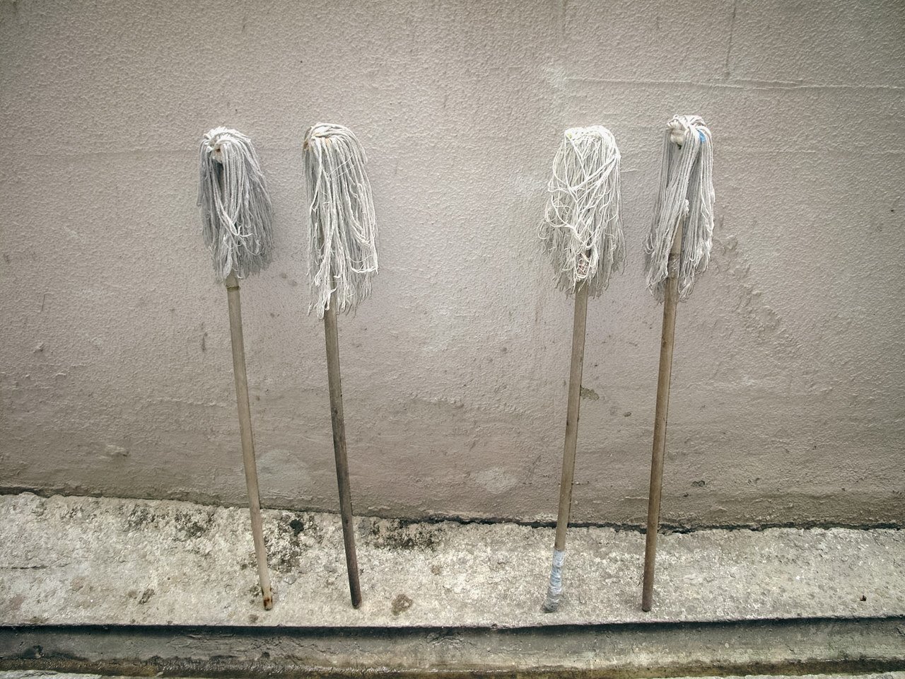Four mop heads with worn white cloths hanging from their handles, leaning against a beige wall.