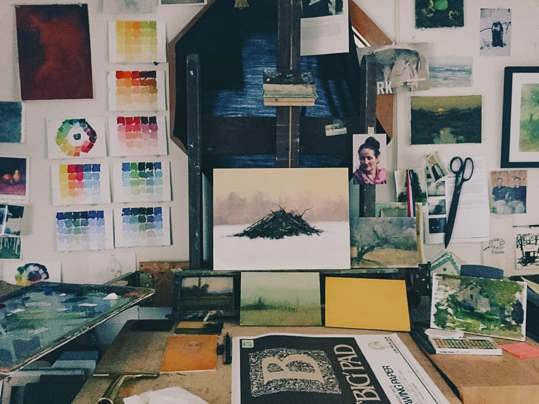 An artist's workspace featuring various paintings, colorful color swatch sheets on the wall, a palette with paint, and art supplies including scissors and brushes.