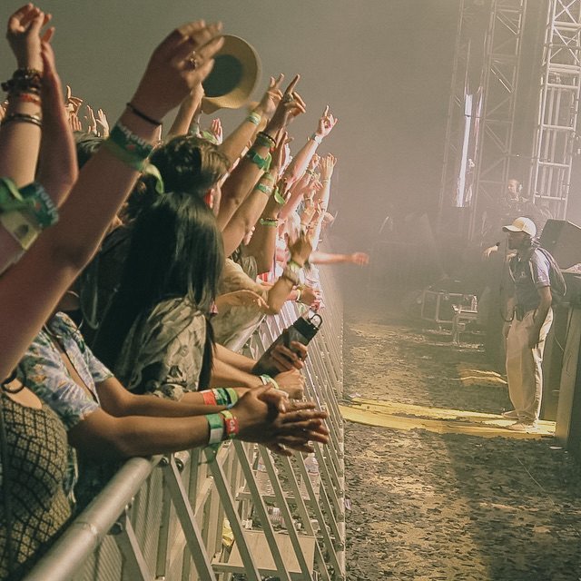 Crowd at a music concert, raising hands and clapping, with security barriers, stage, and stage equipment visible in the background.