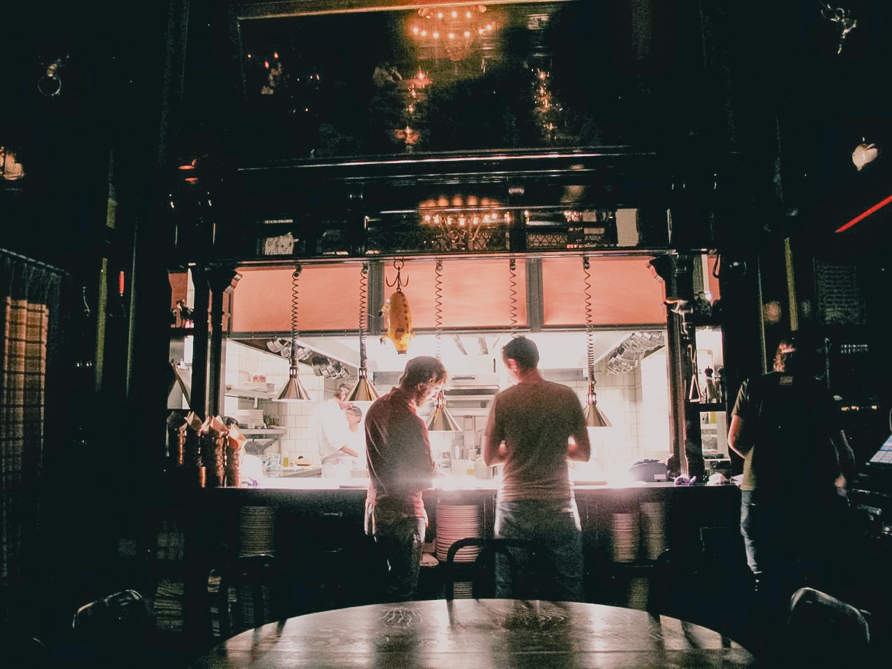 View of a restaurant kitchen seen through an open window, with two chefs and staff preparing food under hanging lights, illuminated by warm lighting inside, and a dark dining area in the foreground.