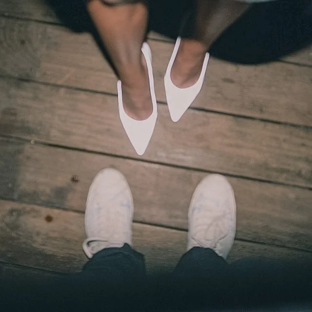 A person wearing white sneakers standing on a wooden floor, viewed from above, with another person wearing white pointed-toe heels also standing on the floor.