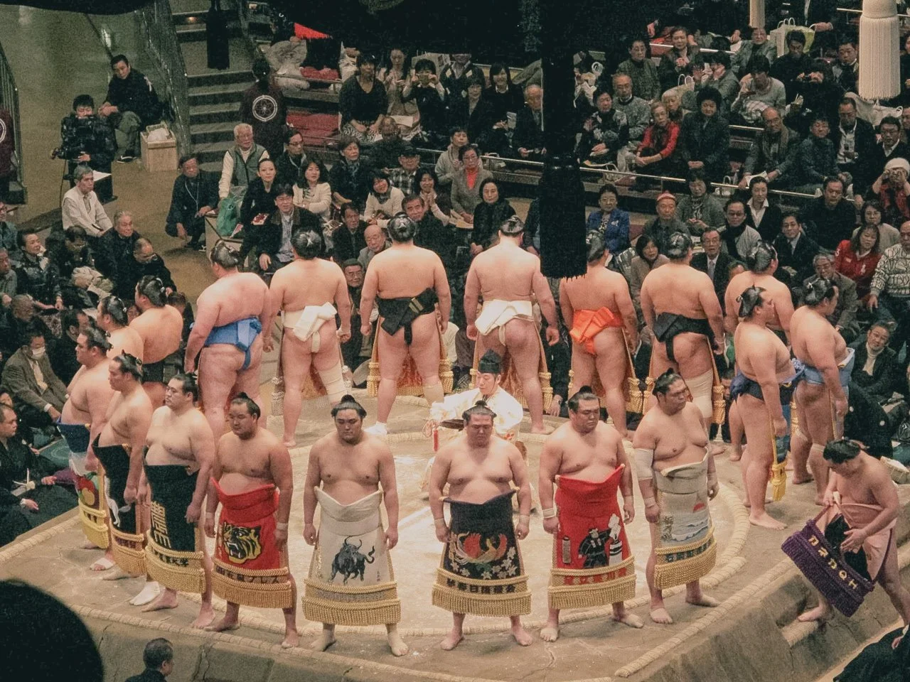 Sumo wrestlers in traditional attire preparing for a match in the ring, with an audience watching attentively.