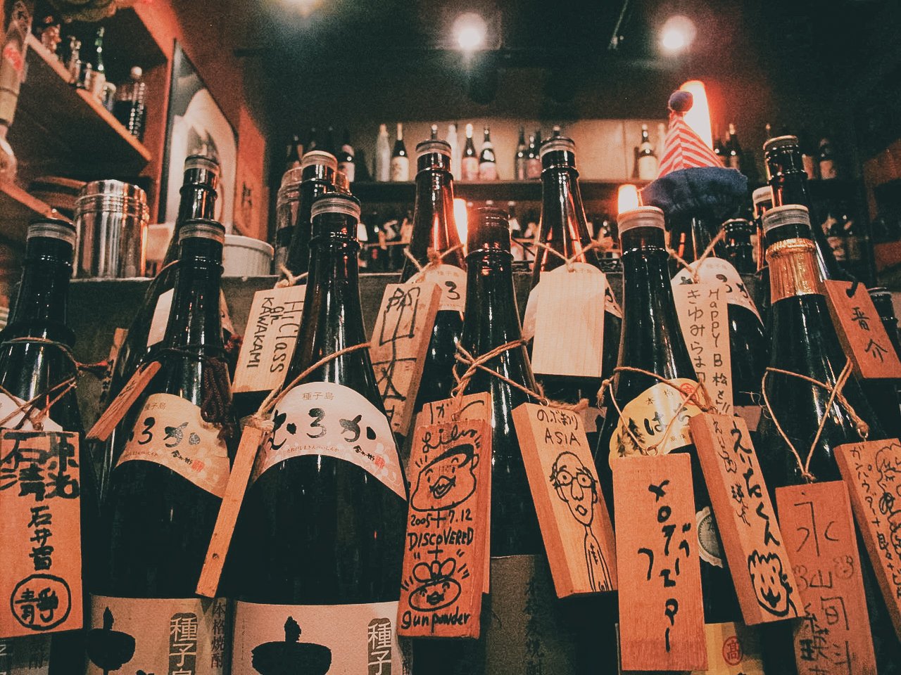Bottles of sake or soju in a bar, with handwritten tags and drawings attached to each bottle.