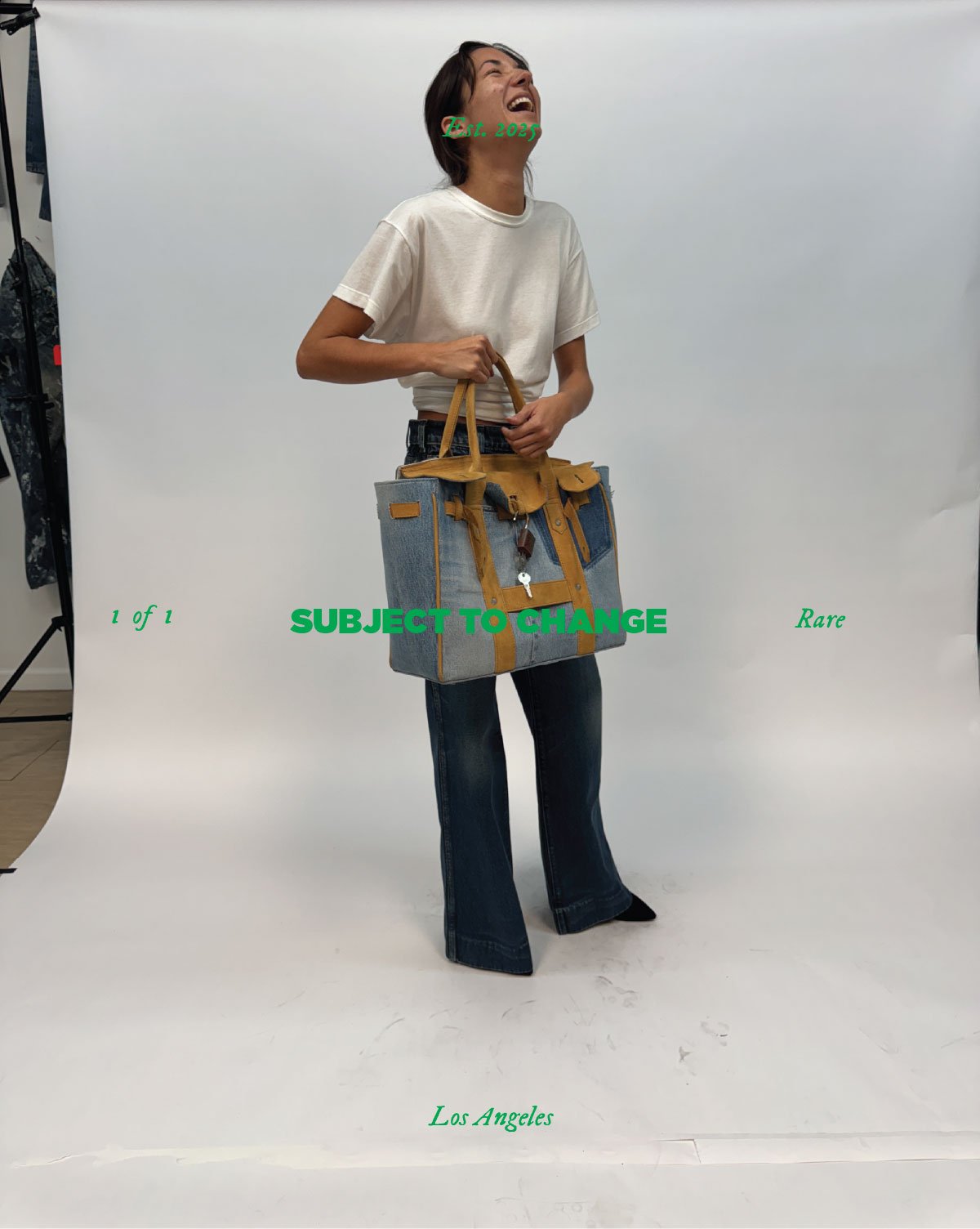 A woman standing with her head thrown back, laughing, holding a large gray and yellow tote bag, in a studio with a white backdrop.
