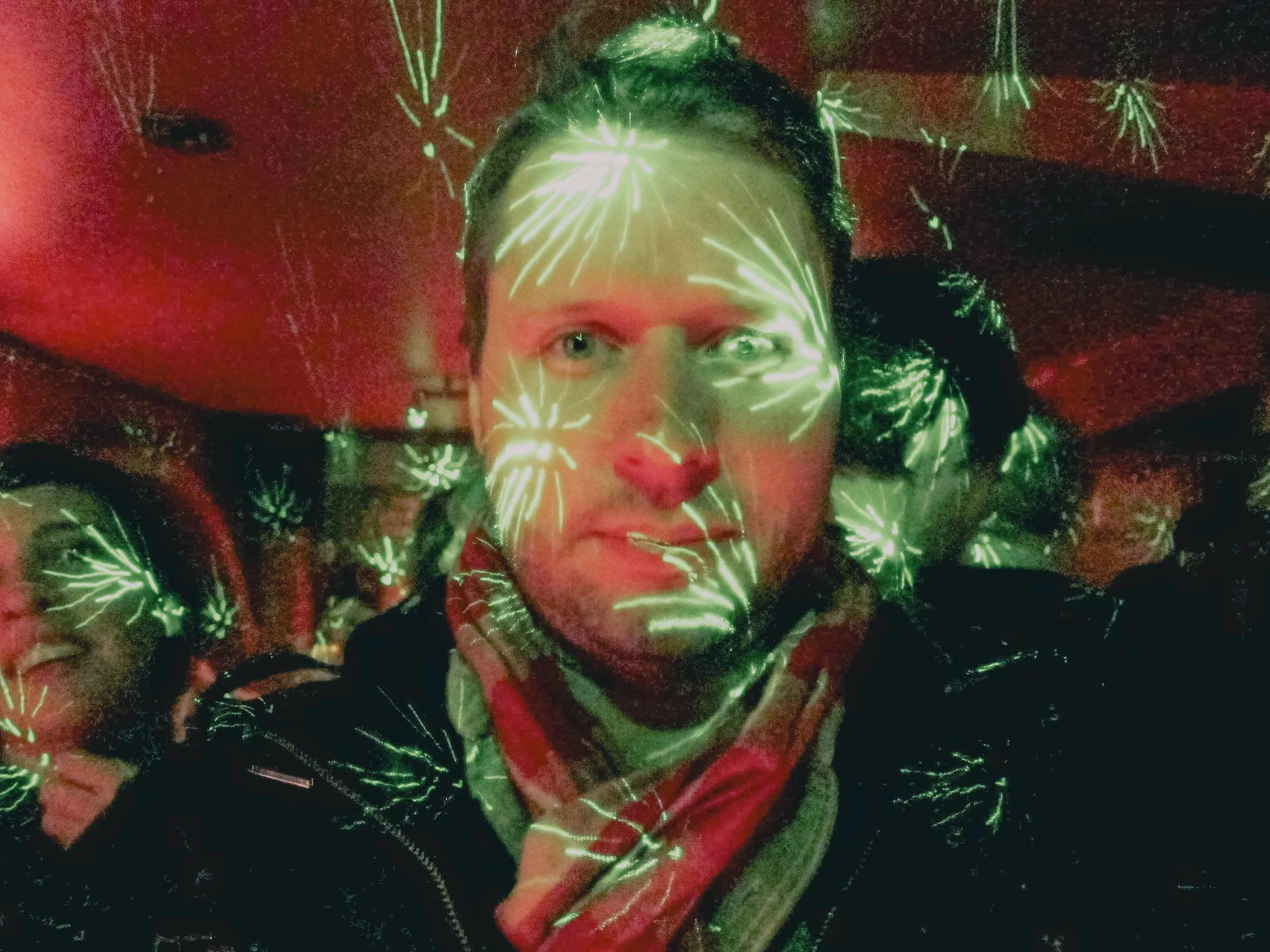 A person with light skin and short hair is surrounded by green laser light reflections that resemble fireworks. The person is looking directly at the camera with a neutral expression, and the background appears to be indoors with dim lighting.